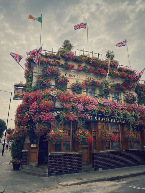 A multi-story pub building with its exterior fully decorated with an abundance of vibrant, flowering plants and hanging baskets, predominantly pink, red, and purple. Several Union Jack flags and the I