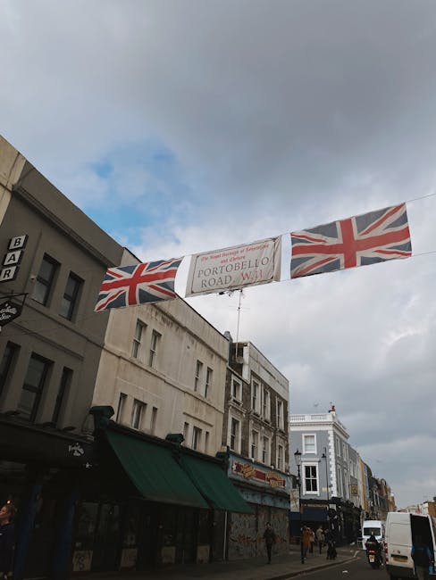 A multi-story pub building with its exterior fully decorated with an abundance of vibrant, flowering plants and hanging baskets, predominantly pink, red, and purple. Several Union Jack flags and the I